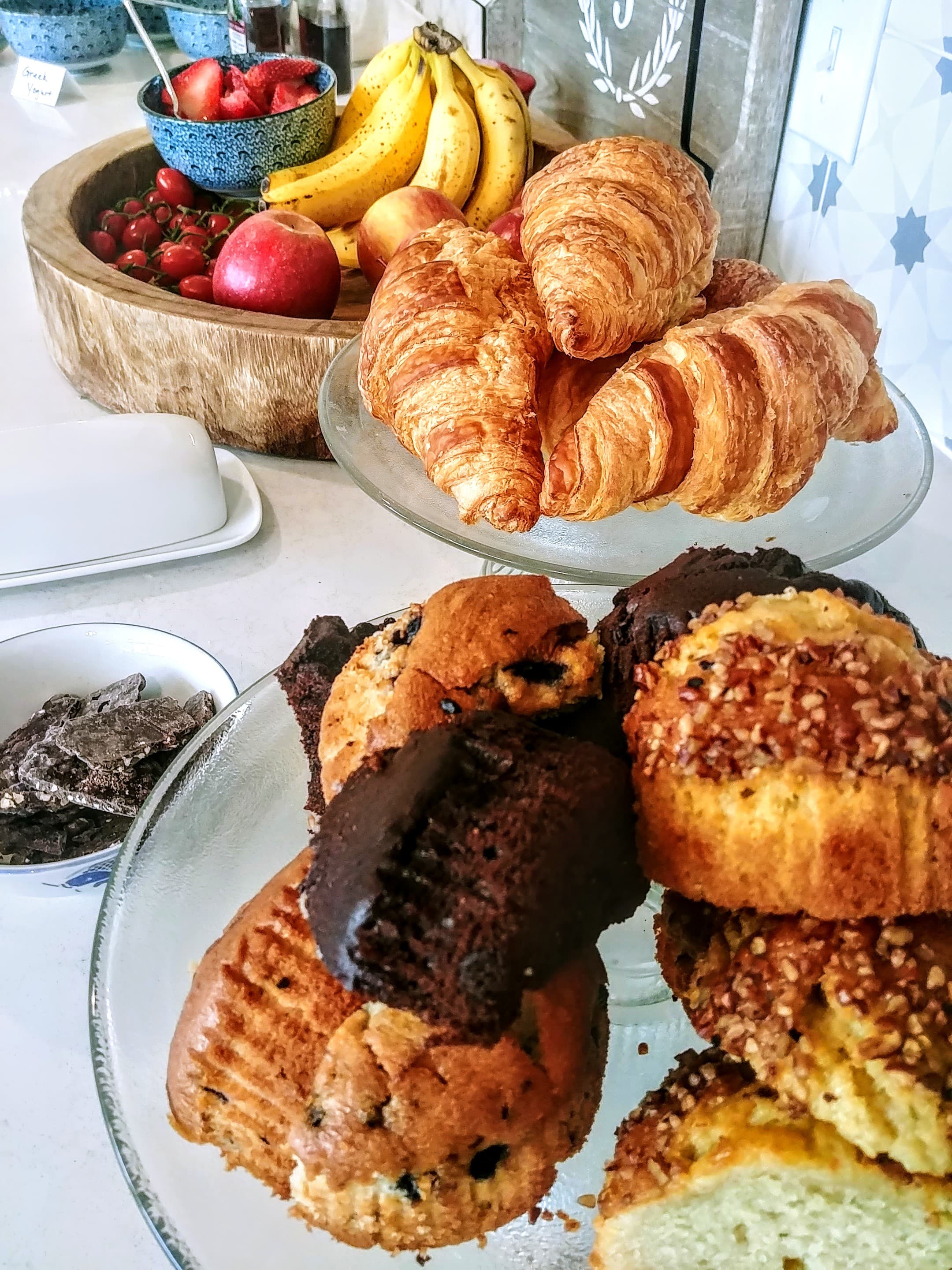 Table set with platters full of freshly baked muffins and croissants and a wooden bowl of fresh fruit including bananas, strawberries, and apples