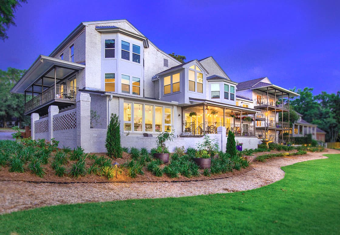 A spacious, two-story house with a white facade and multiple windows. The house features a wraparound porch and a landscaped backyard with a gravel path winding through lush greenery. The scene is illuminated by warm evening light.