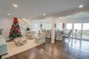 A spacious living area featuring a decorated Christmas tree, light-colored furniture, and a dining area with large windows.