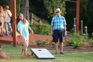 A woman throws a bean bag toward a cornhole board while a man stands nearby watching.