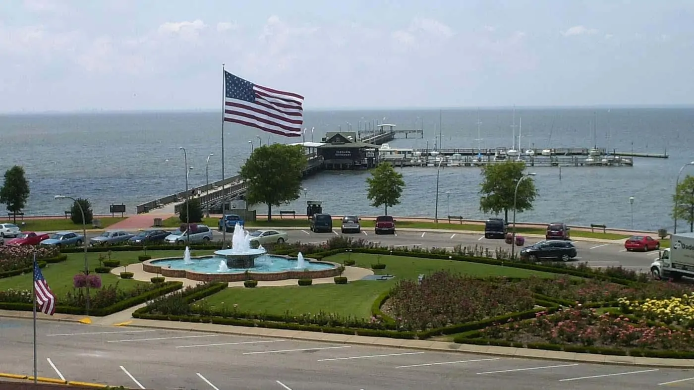 A scenic view of a waterfront area featuring a fountain, American flag, and marina in the background.